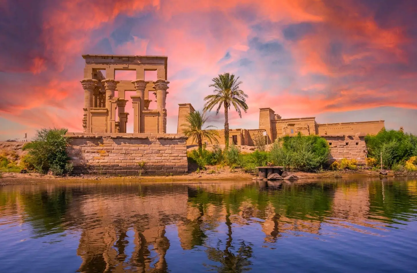 The ancient Philae Temple complex on Agilkia Island in Aswan, Egypt, reflecting on the calm waters of the Nile River under a vibrant sunset sky. Featured in the Aswan Egypt Ultimate Guide.