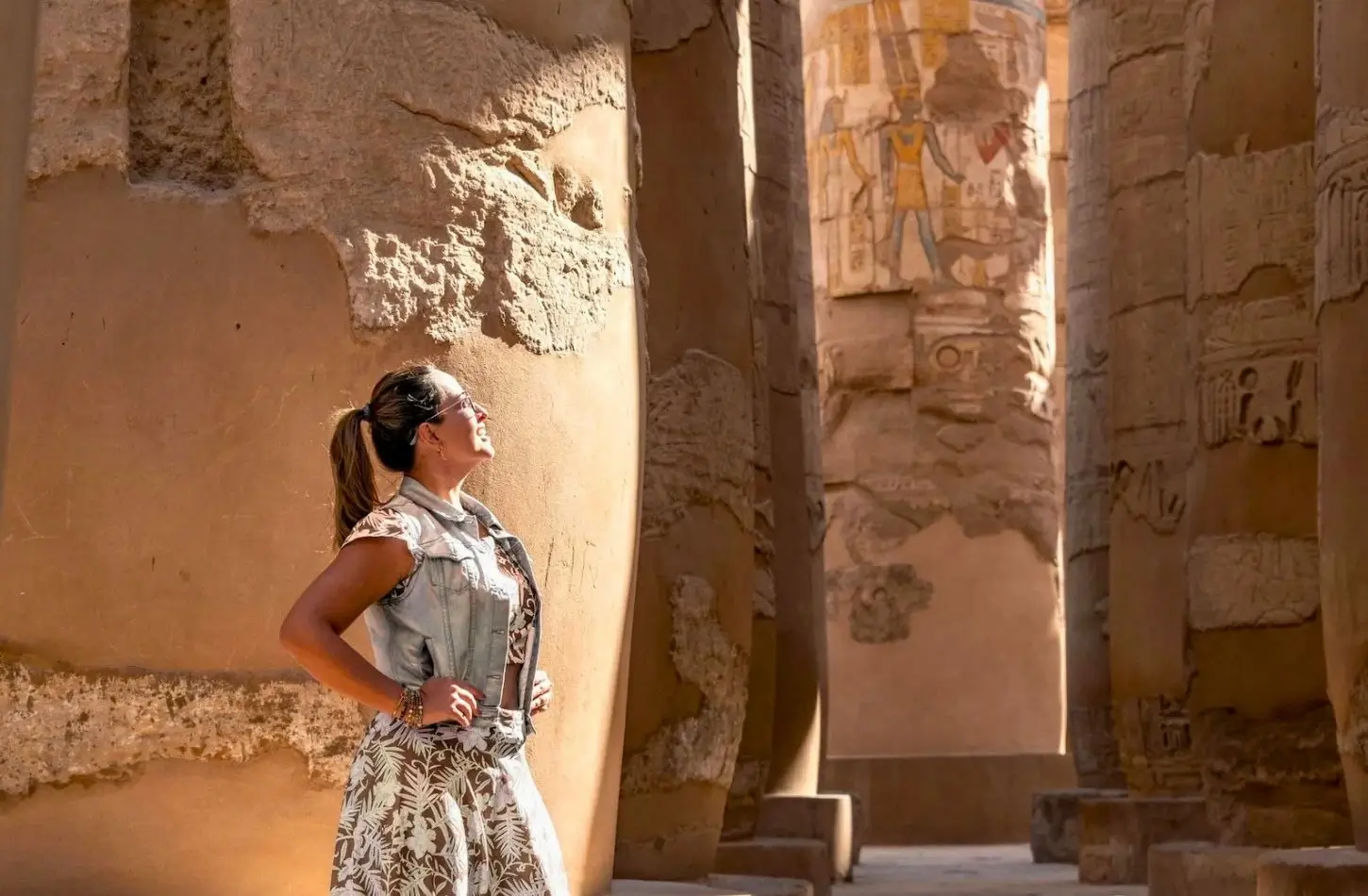 A woman standing among the massive ancient columns of Karnak Temple in the City of Luxor, Egypt, admiring the detailed hieroglyphics and carvings under bright sunlight.