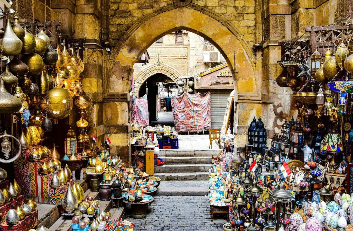 A bustling market stall in Cairo's Khan El-Khalili bazaar filled with hanging brass lamps, mosaic lanterns, and Egyptian souvenirs under a historic stone archway, perfect for a day trip from Marsa Alam