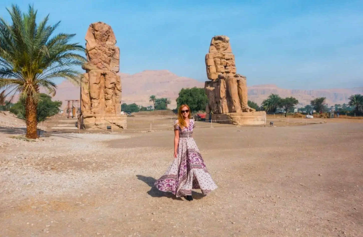 Nejlepší časy k návštěvě Egypta ukazují jasnou oblohu u Kolosů Memnona v Luxoru, kde stojí turista v maxi šatech před obrovskými starobylými kamennými sochami.