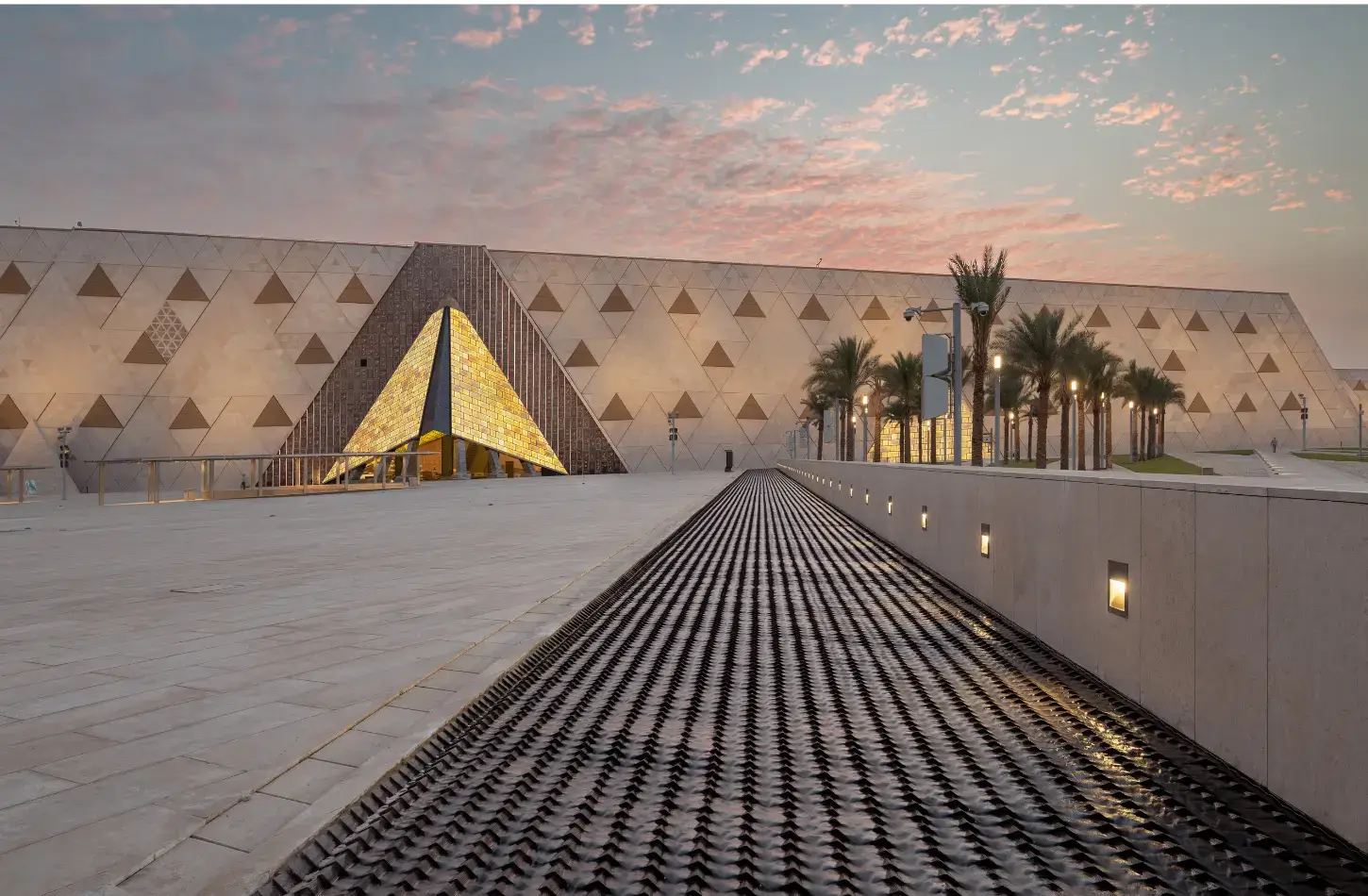 Facciata esterna del Grande Museo Egiziano GEM al tramonto, che mostra il moderno muro di pietra traslucida con motivi triangolari, un ingresso a piramide dorata illuminata e palme che fiancheggiano la piazza principale.