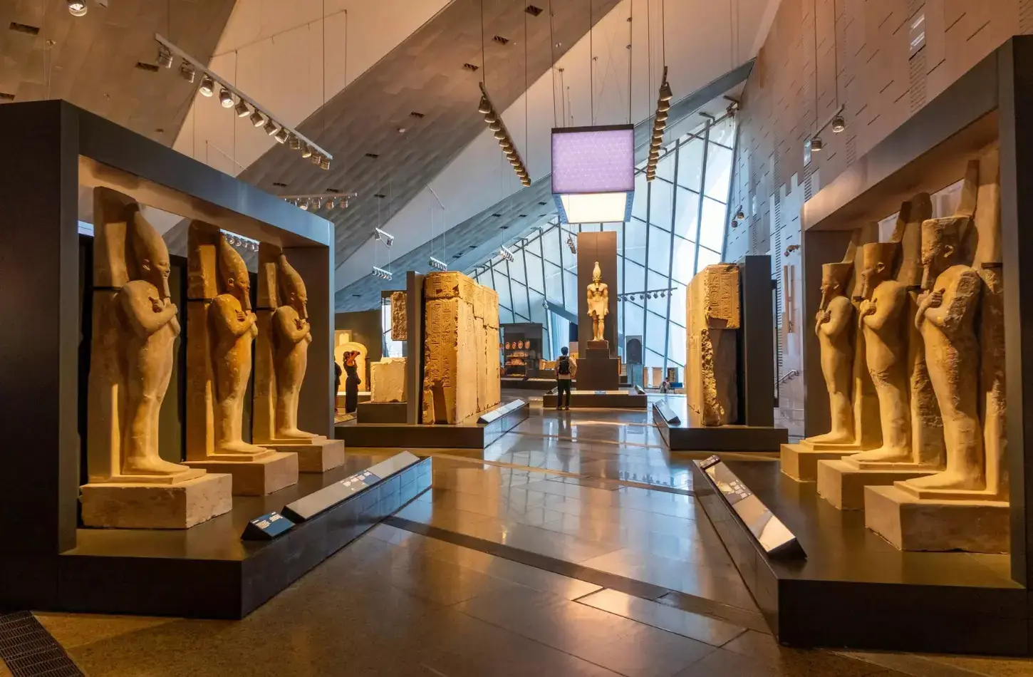 Vista interna delle gallerie principali ultra-moderne del Grande Museo Egiziano GEM a Giza, con file di statue monumentali di antichi faraoni, pareti di calcare con geroglifici e una facciata di vetro durante un tour storico guidato.