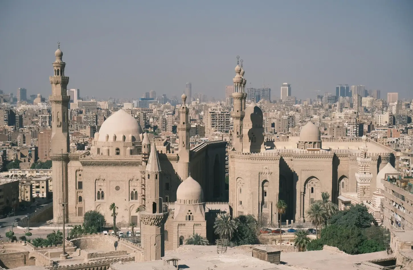 A high-angle panoramic view of the historic Sultan Hassan and Al-Rifa'i mosques in Islamic Cairo, showcasing their massive domes and soaring minarets against the sprawling city skyline, ideal for a day excursion 