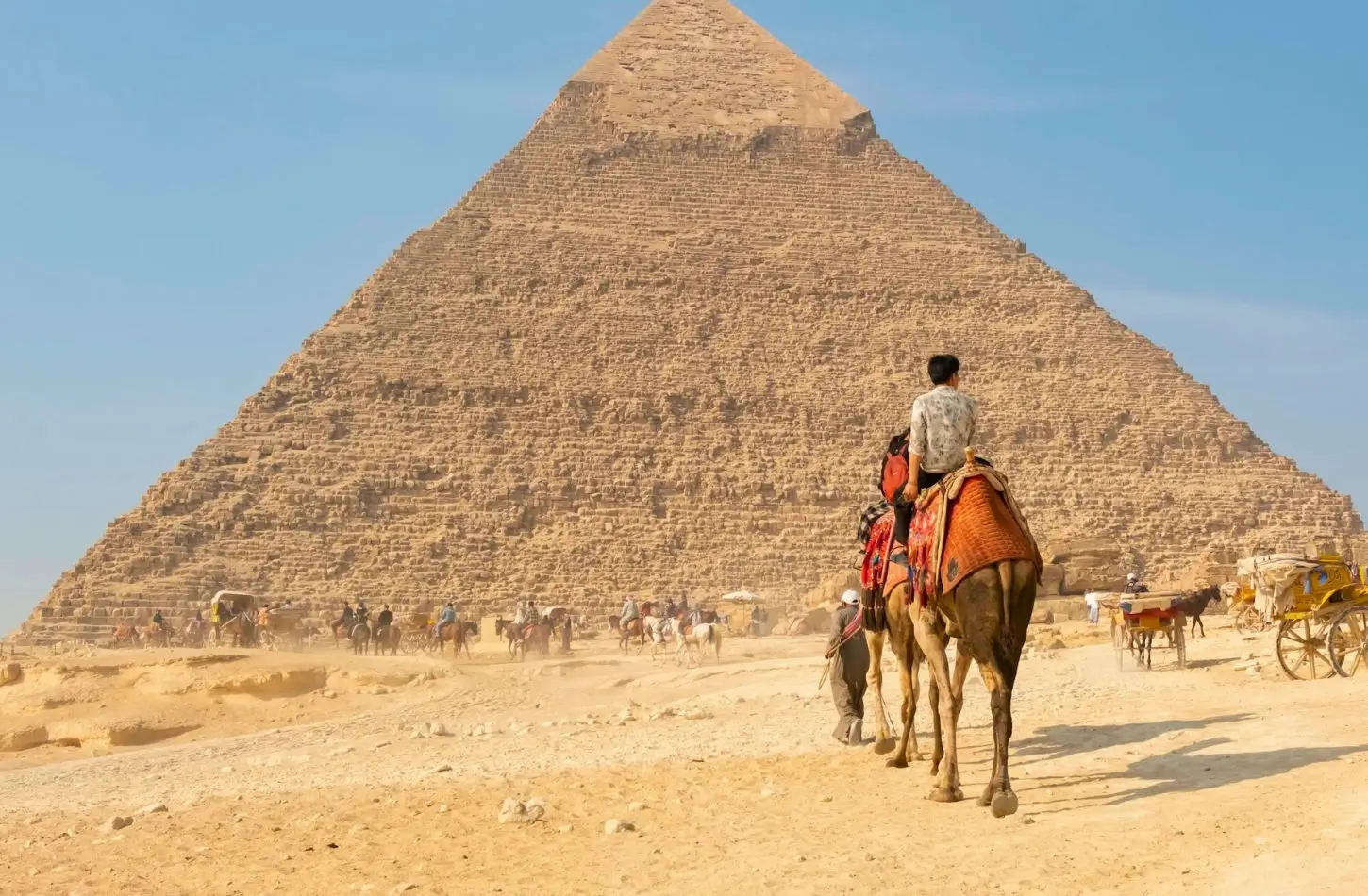 A tourist riding a camel across the desert sands in front of the massive Pyramid of Khafre, one of the famous Pyramids of Giza, under a clear blue sky.