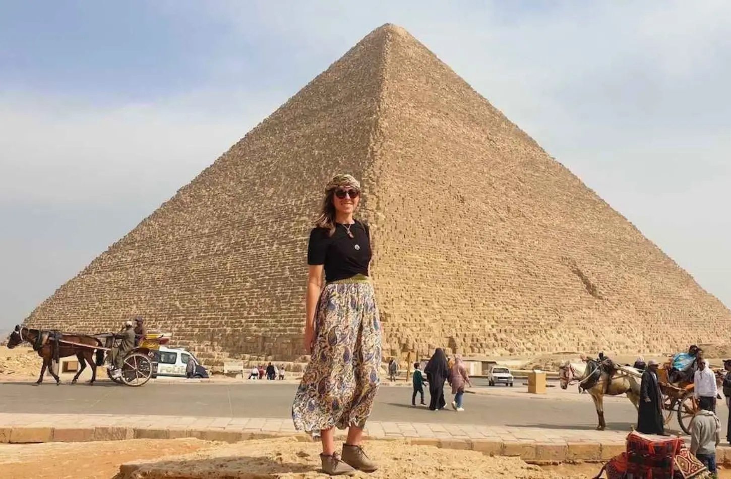 Žena stojící před Velkou pyramidou v Gíze, jedním z nejlepších míst k návštěvě v Egyptě, s koňskými povozy a turisty v pozadí.